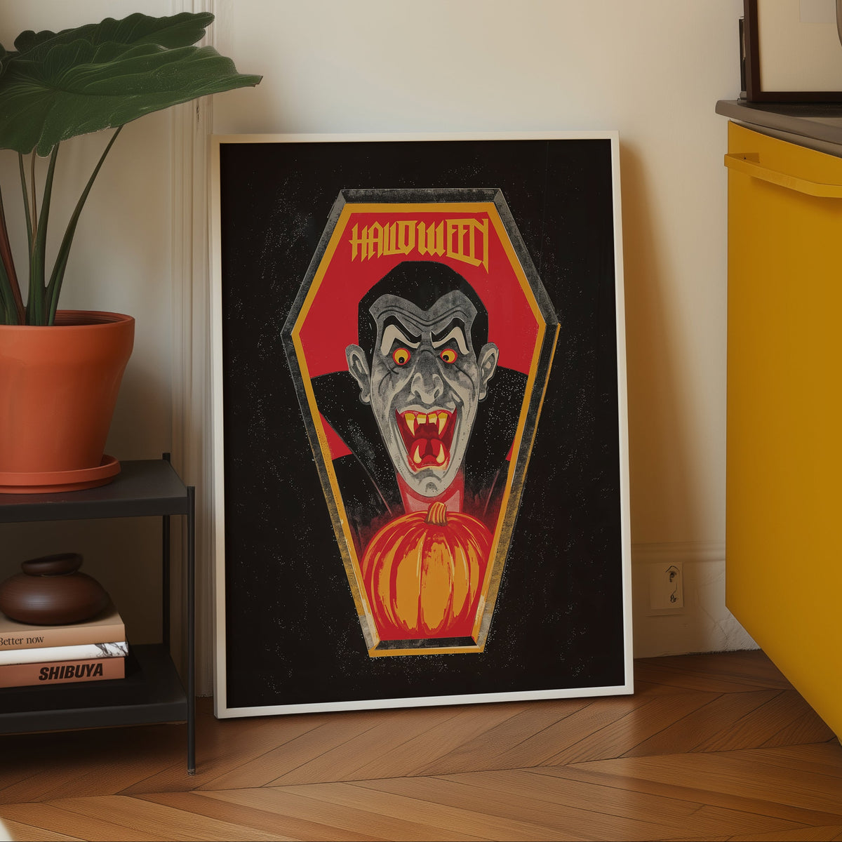 A framed gothic Halloween art print featuring a vampire in a coffin with 'HALLOWEEN' text, leaning against a wall in a modern kitchen setting next to a yellow cabinet and a potted plant.