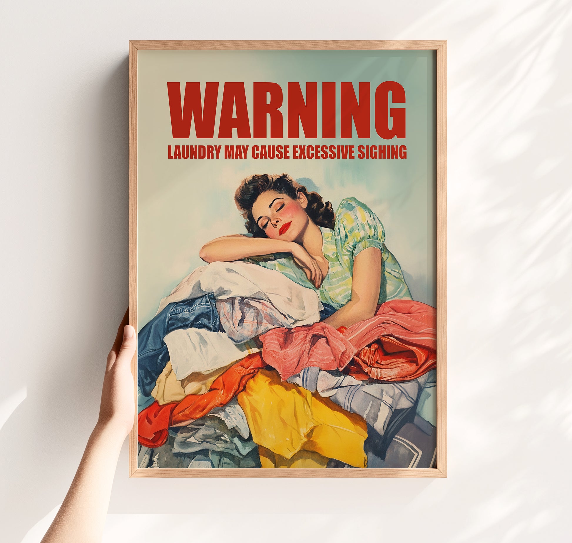 Vintage illustration of a 1950s housewife leaning her head on a large pile of colorful laundry with 'WARNING: LAUNDRY MAY CAUSE EXCESSIVE SIGHING' in bold red text; funny retro laundry room wall art.