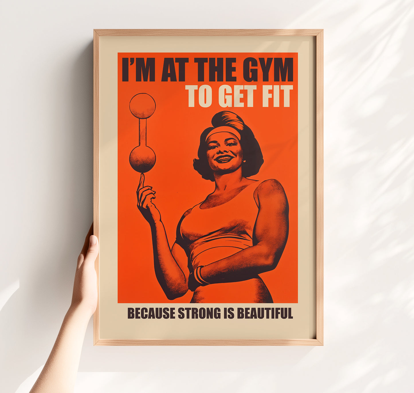 Vintage-style gym poster on an orange background featuring a muscular woman balancing a barbell on one finger with the text "I'M AT THE GYM TO GET FIT BECAUSE STRONG IS BEAUTIFUL."