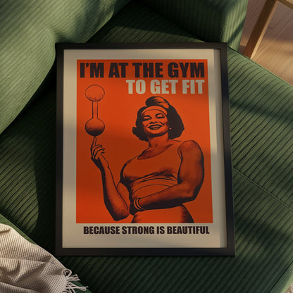Vintage-style gym poster on an orange background featuring a muscular woman balancing a barbell on one finger with the text "I'M AT THE GYM TO GET FIT BECAUSE STRONG IS BEAUTIFUL."
