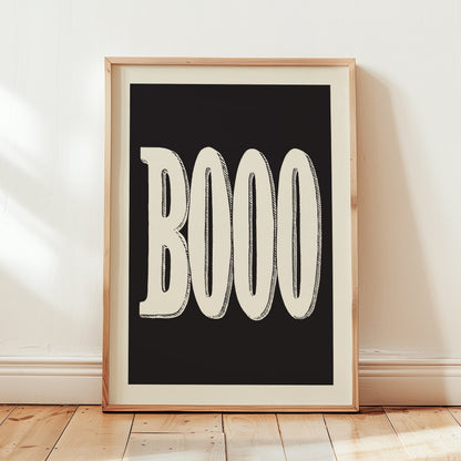 A framed typographic art print leaning against a white wall on a light wood floor. The print features the word 'BOOO' in large cream-colored letters with hand-drawn black outlines on a solid black background, enclosed in a light natural wood frame.