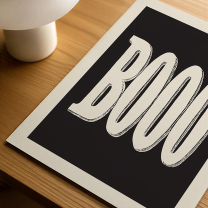 A close-up of a typographic art print featuring the word 'BOO' in large white outlined letters on a black background, photographed at an angle on a wooden surface with part of a white lamp visible.