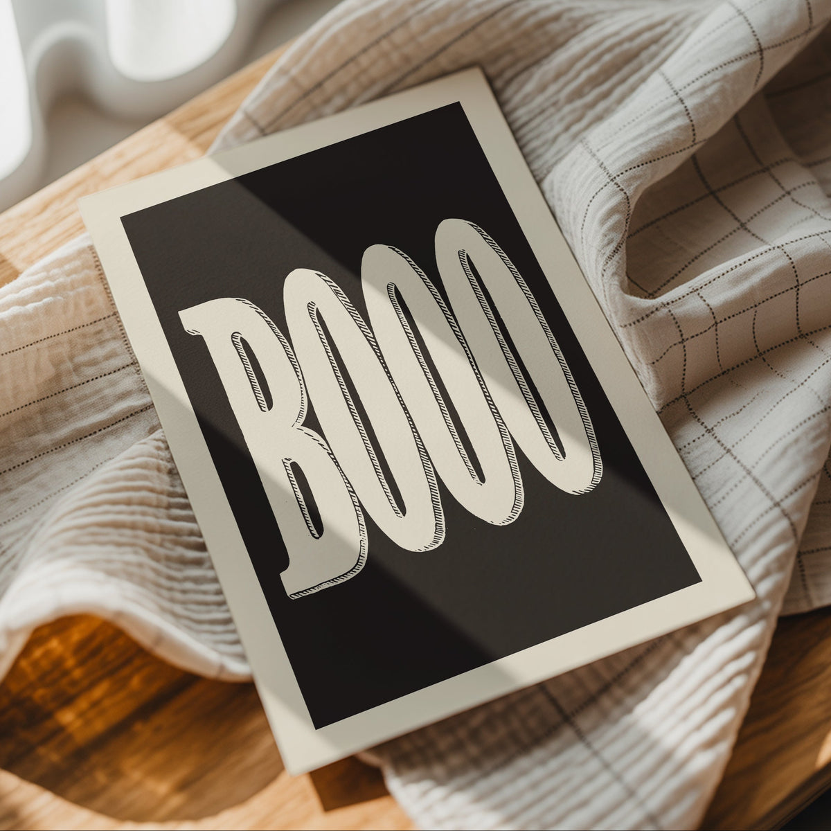 A square art print with the word 'BOO' in large white stylized letters on a black background, styled on a beige plaid cloth over a wooden surface.