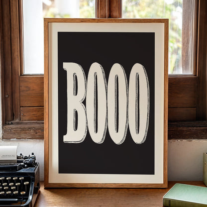 A framed typographic art print featuring the word 'BOOO' in large cream letters on a black background, leaning on a wooden table near a window with a vintage typewriter, books, and a candle nearby.