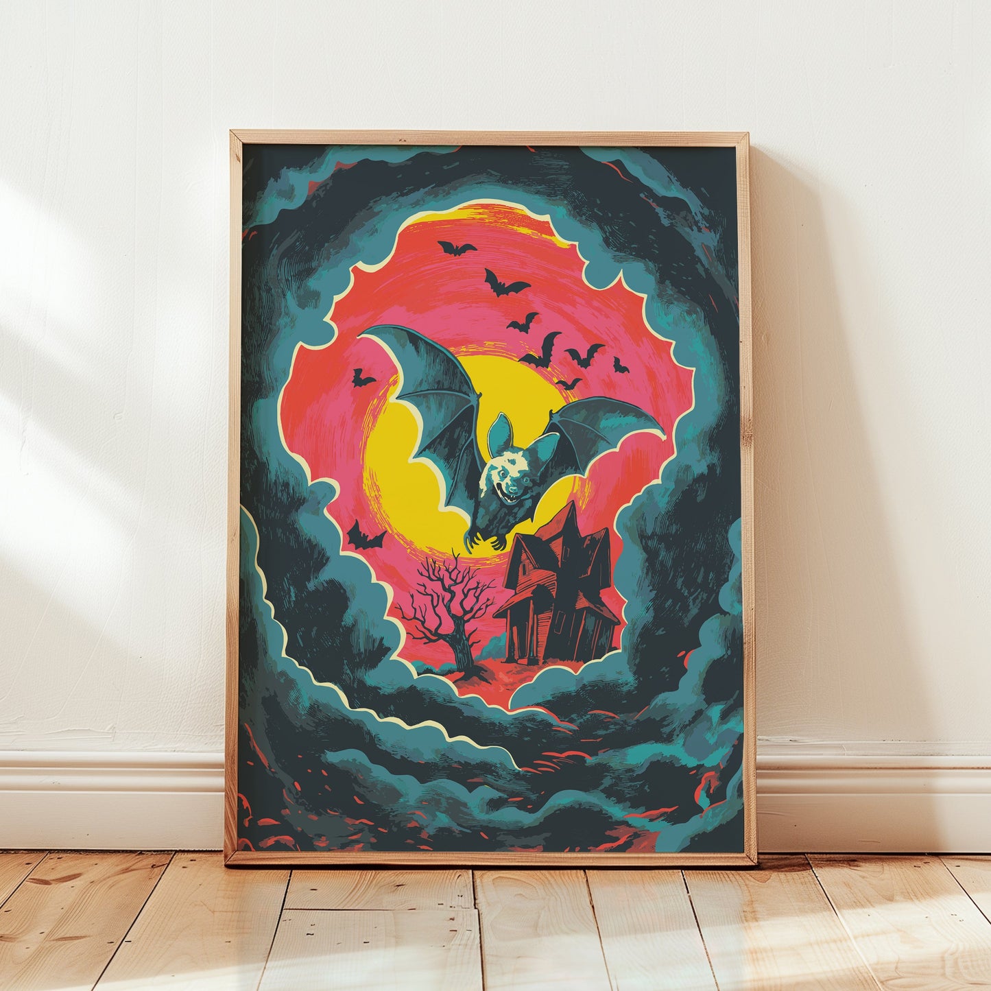 A framed art print featuring a whimsical illustration of a large bat flying over a haunted house, set against a swirling pink, yellow, and teal sky. The print is displayed leaning against a white wall on a light wooden floor.