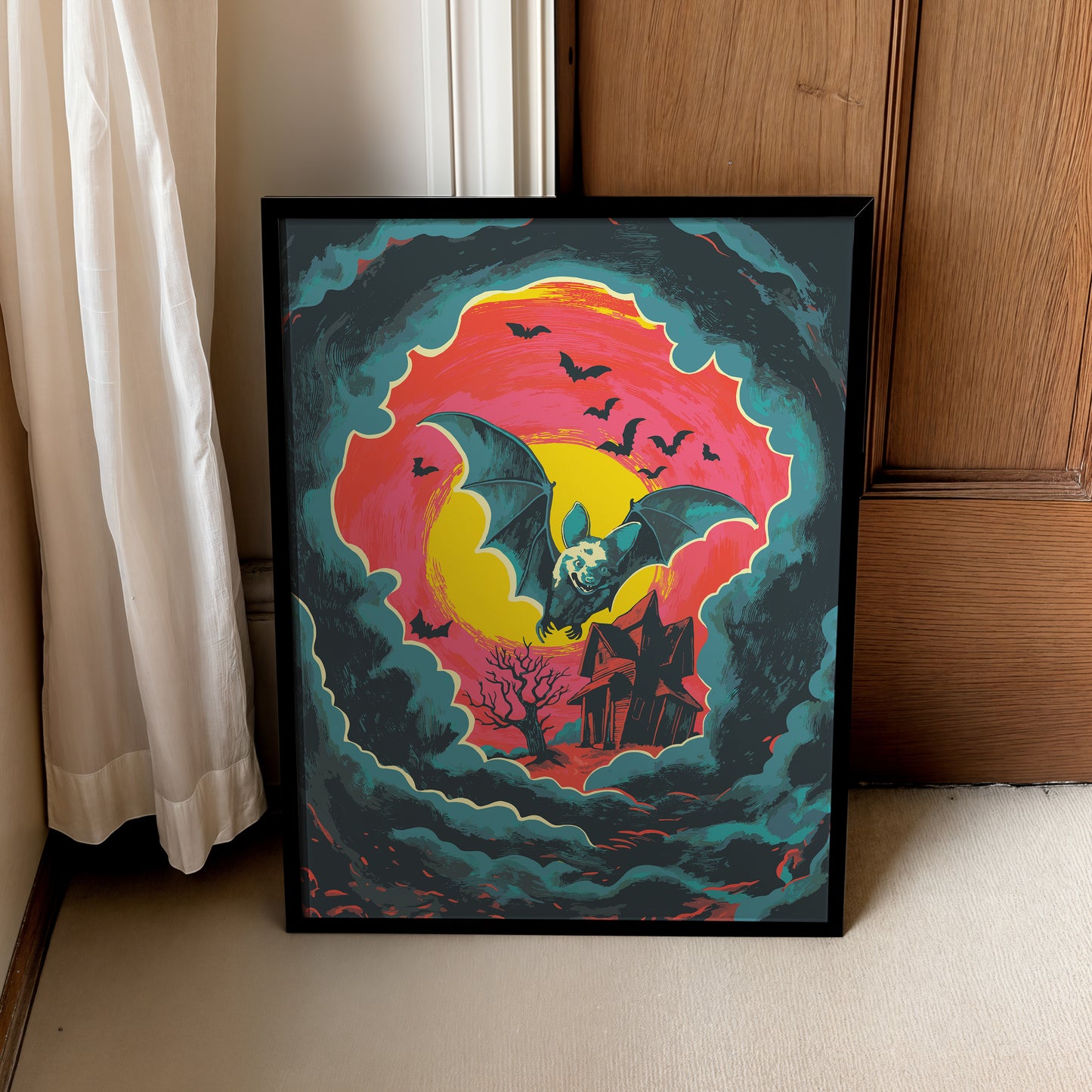 A framed art print leaning against a wooden door and white curtain on a beige floor. The artwork features a bat flying in front of a bright yellow and pink sun, surrounded by swirling teal clouds, with a haunted house and bare tree below.
