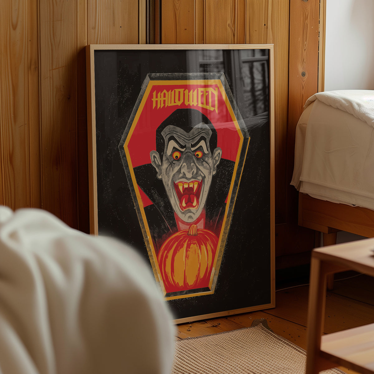 A framed Halloween art print featuring a vampire inside a coffin with a pumpkin, leaning against a wooden wall in a bedroom setting.