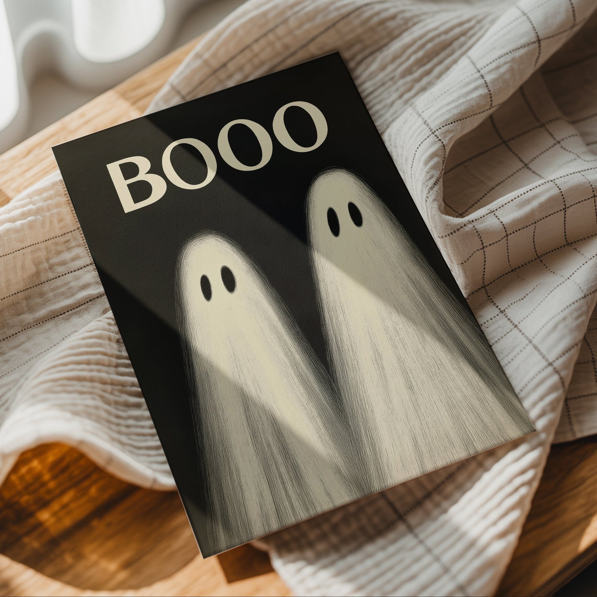 A square art print with a black background featuring the word 'BOOO' in cream and two ghostly figures, styled on a beige checkered cloth over a wooden surface.