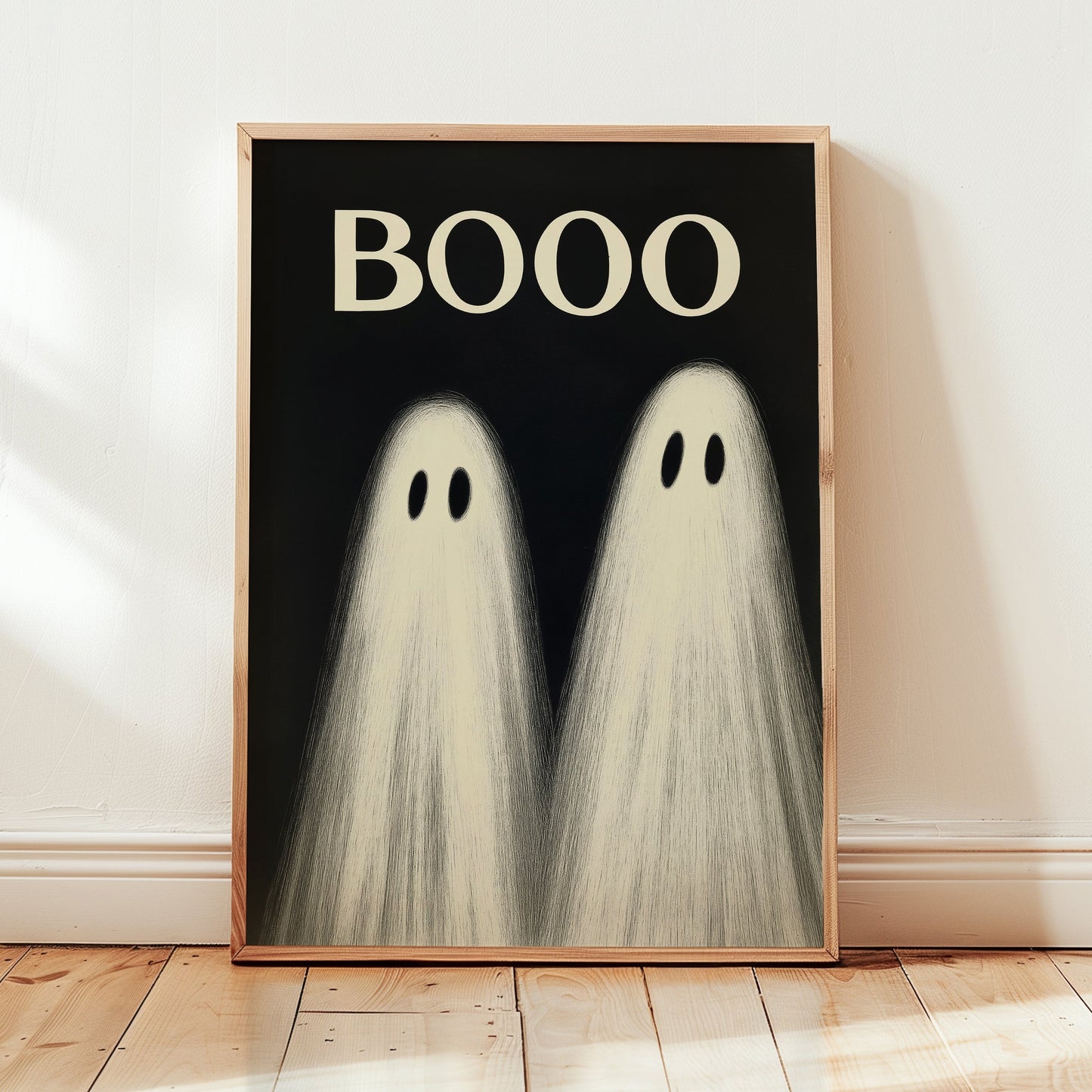 A framed art print featuring two cute cartoon ghosts with the word 'BOOO' in cream text on a black background, leaning against a white wall on a light wood floor.