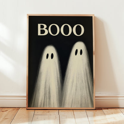 A framed art print featuring two cute cartoon ghosts with the word 'BOOO' in cream text on a black background, leaning against a white wall on a light wood floor.