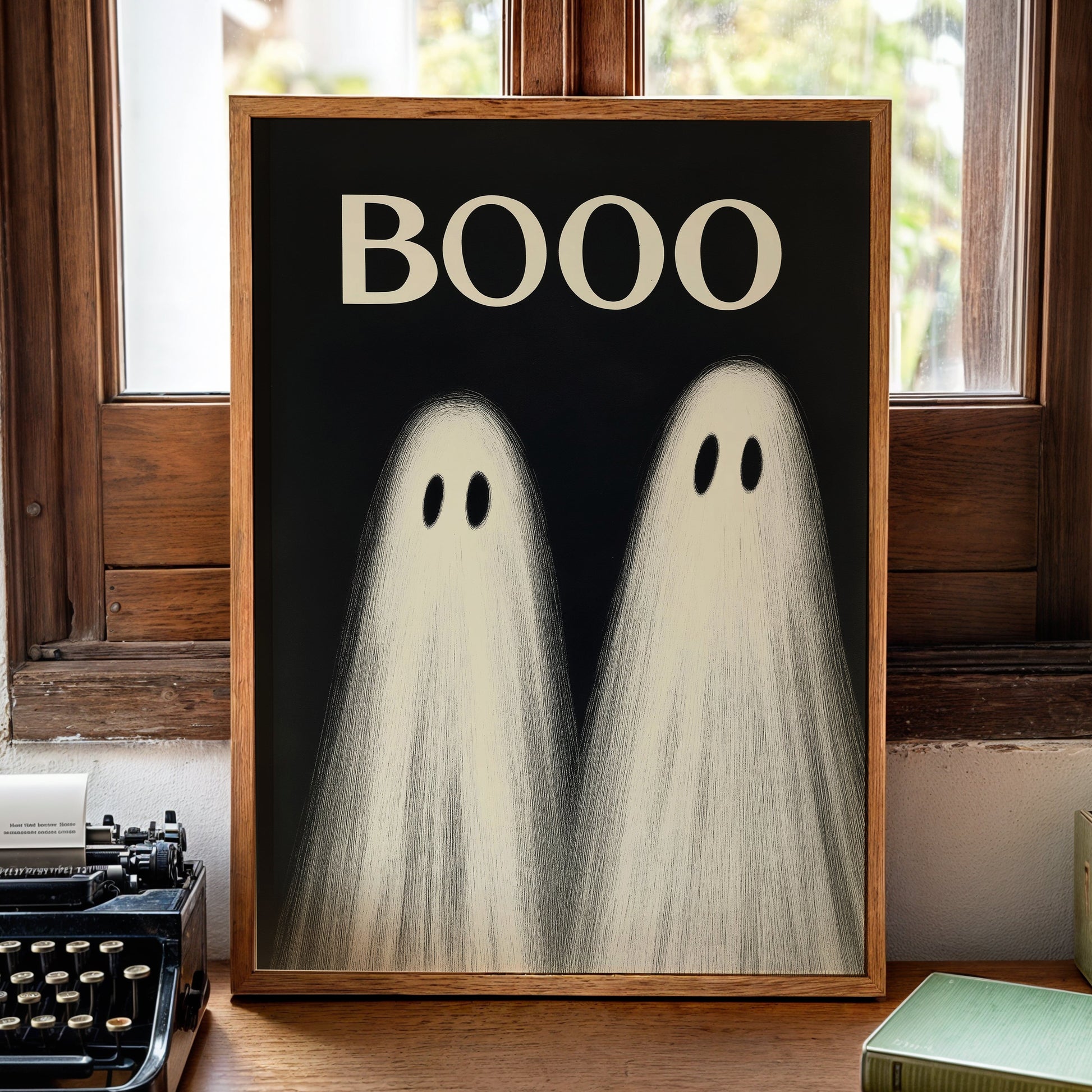 A framed art print featuring two ghostly figures with the word 'BOOO' above them, displayed on a wooden surface in front of a window.