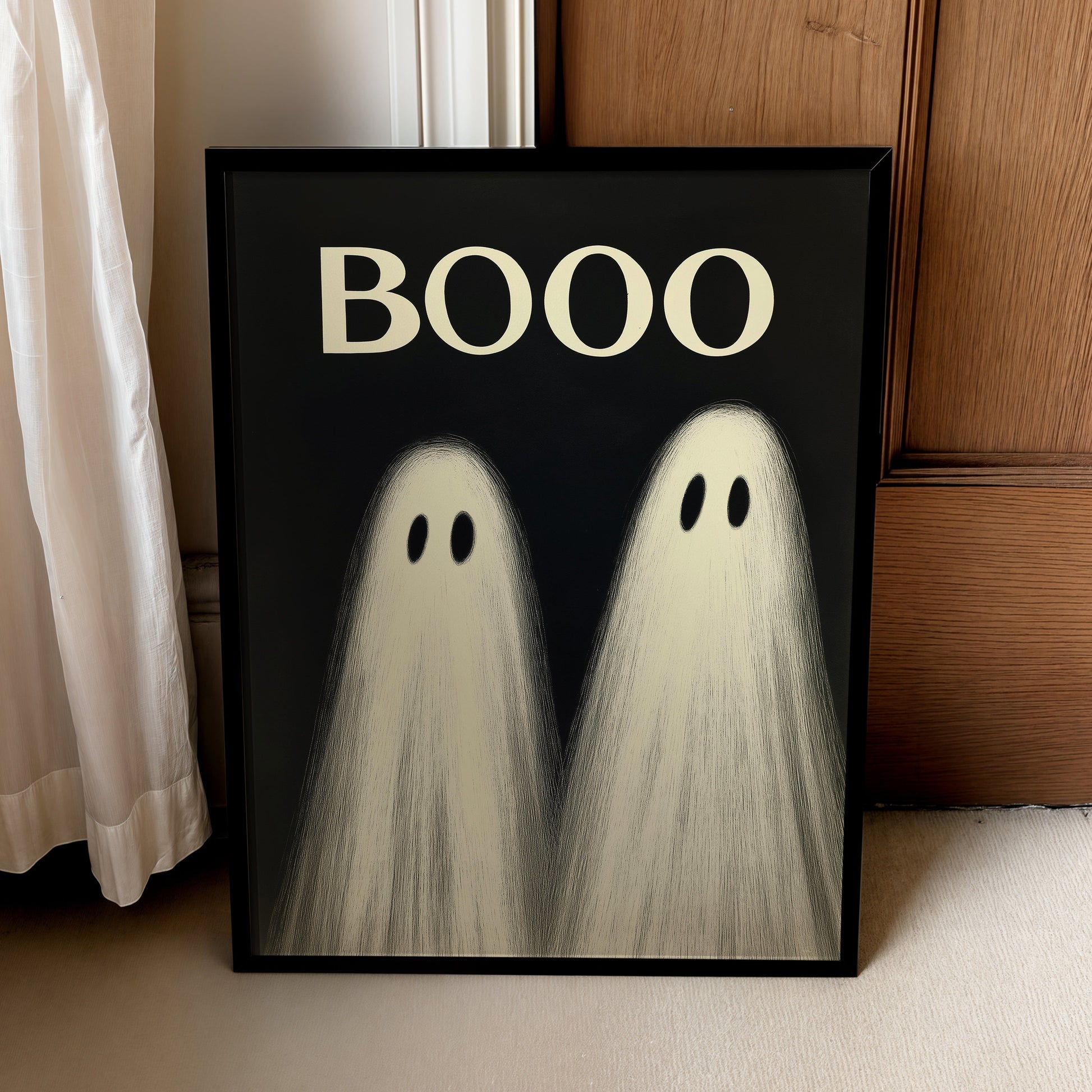 A framed Halloween art print leaning against a wooden door and curtain. The black-framed poster features the word 'BOOO' in cream at the top, above two ghost figures with black eyes on a black background.