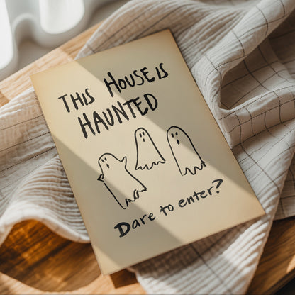 A square cream-colored art print with hand-drawn black text reading 'THIS HOUSE IS HAUNTED' and 'Dare to enter?' with three simple ghost illustrations, styled on a wooden surface with a checkered cloth.