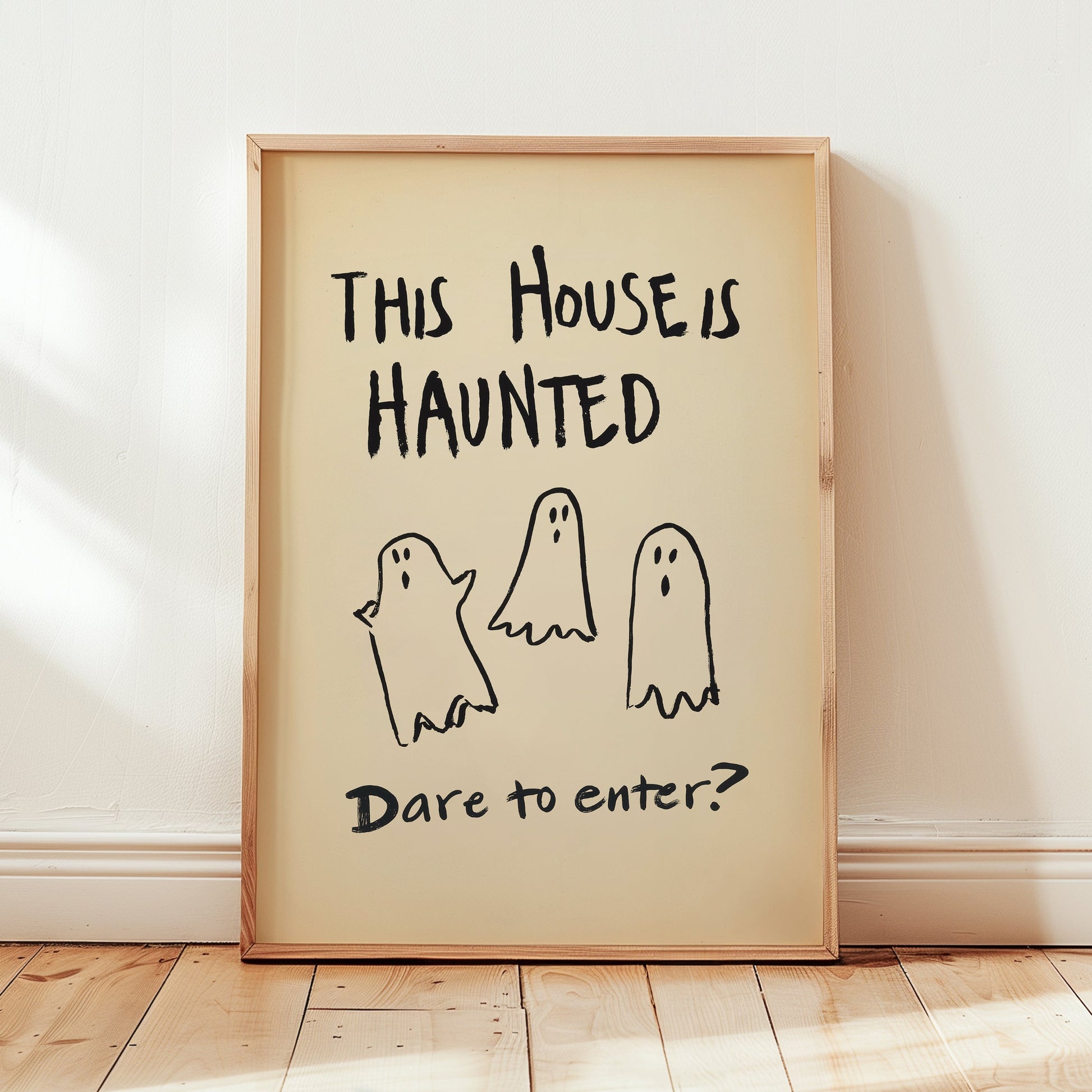 A framed Halloween art print leaning against a white wall on a light wood floor. The print features the text 'THIS HOUSE IS HAUNTED' and 'Dare to enter?' with three simple black ghost illustrations on a cream background, enclosed in a light natural wood frame.