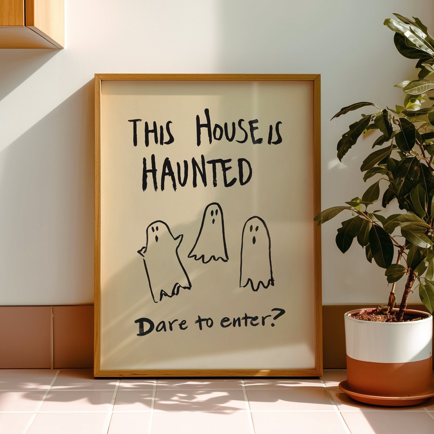 A framed art print with the text 'THIS HOUSE IS HAUNTED' and 'Dare to enter?' with three simple ghost drawings, leaning against a wall in a sunlit room next to a potted plant.