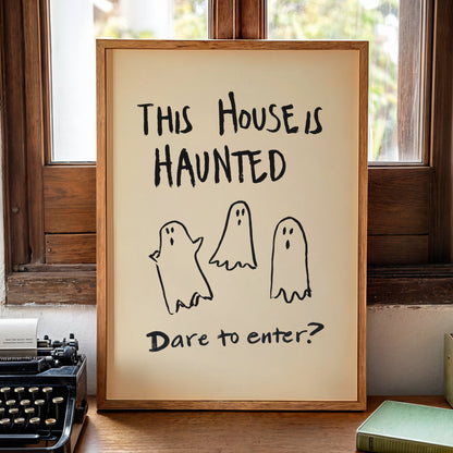 A framed art print with the text 'THIS HOUSE IS HAUNTED' and 'Dare to enter?' with three simple ghost illustrations, displayed on a wooden table in a cozy room setting next to a typewriter and candle.