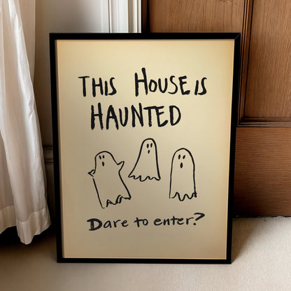 A framed Halloween art print leaning against a wooden door and white curtain. The print features a beige background with black text reading 'THIS HOUSE IS HAUNTED' and 'Dare to enter?' along with three simple hand-drawn ghost illustrations.
