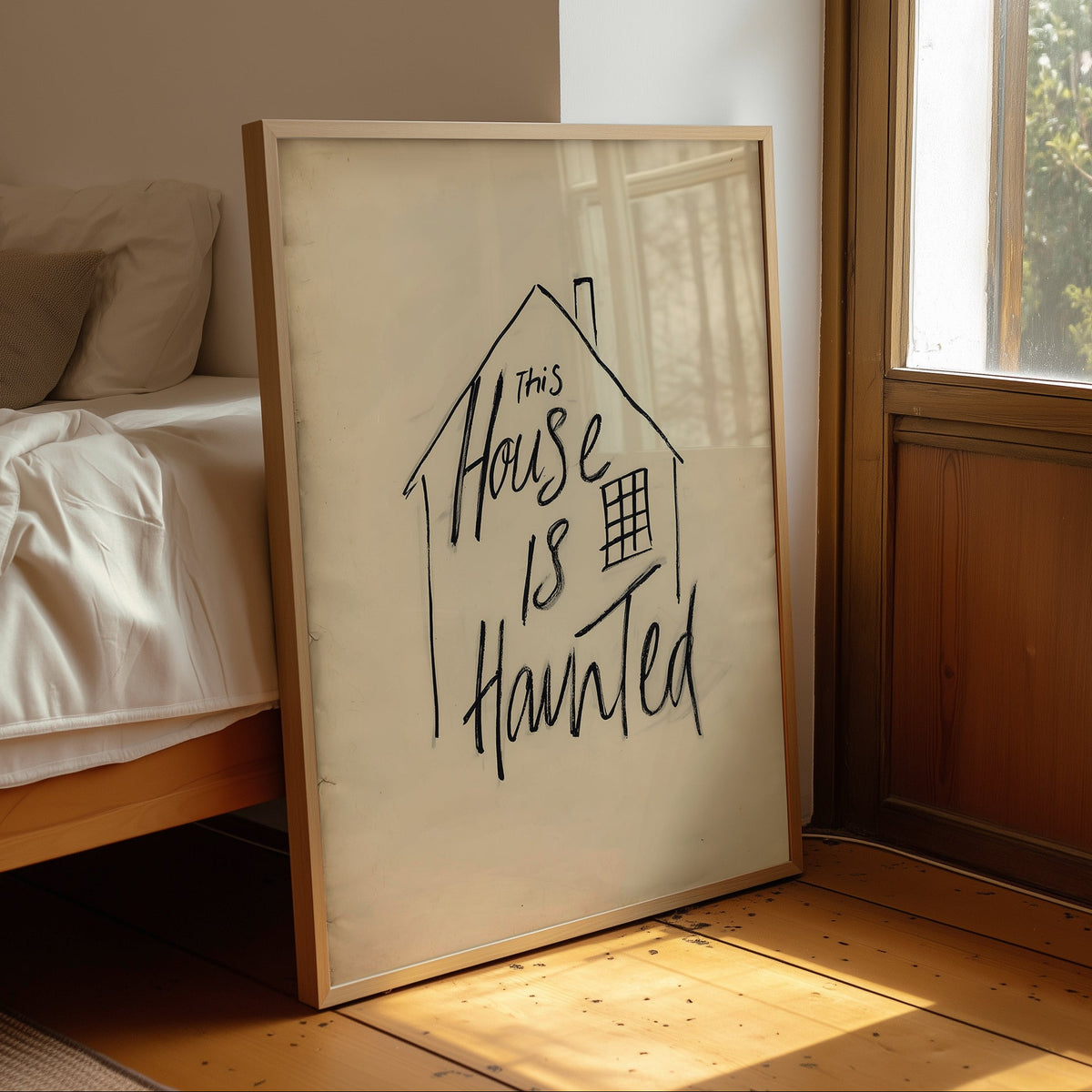 A framed art print leaning against a bed in a sunlit bedroom. The print features a simple house outline with the handwritten phrase 'This House Is Haunted' in black ink on an off-white background, enclosed in a light wood frame.