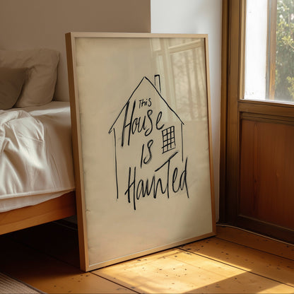 A framed art print leaning against a bed in a sunlit bedroom. The print features a simple house outline with the handwritten phrase 'This House Is Haunted' in black ink on an off-white background, enclosed in a light wood frame.