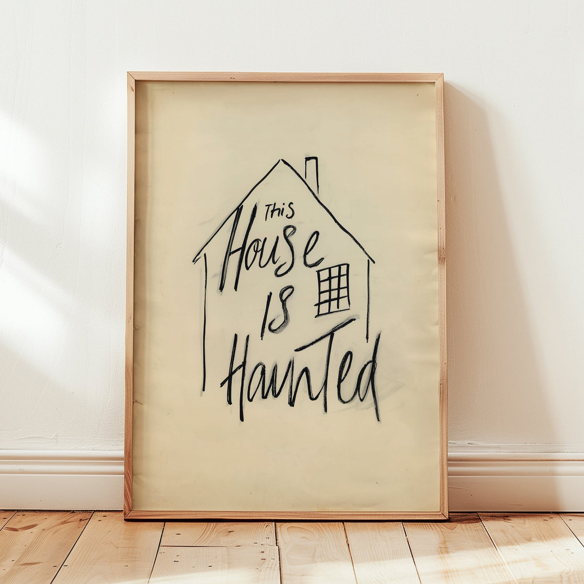 A framed art print leaning against a white wall on a light wood floor. The print features a hand-drawn black ink house outline with the phrase 'This House Is Haunted' written inside on a warm cream background, enclosed in a light natural wood frame.