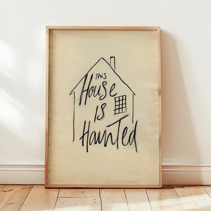 A framed art print leaning against a white wall on a light wood floor. The print features a hand-drawn black ink house outline with the phrase 'This House Is Haunted' written inside on a warm cream background, enclosed in a light natural wood frame.