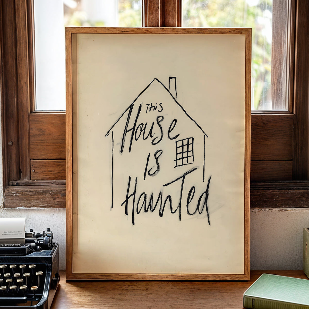 A framed art print with the handwritten text 'This House Is Haunted' in black ink on a cream background, displayed on a wooden desk in a cozy room setting next to a vintage typewriter, books, and a candle.