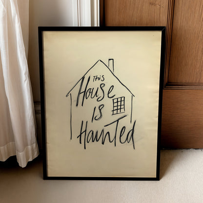 A framed art print with a hand-drawn black ink house outline and the text 'This House Is Haunted' on a cream-colored background, leaning against a wooden door and white curtain.