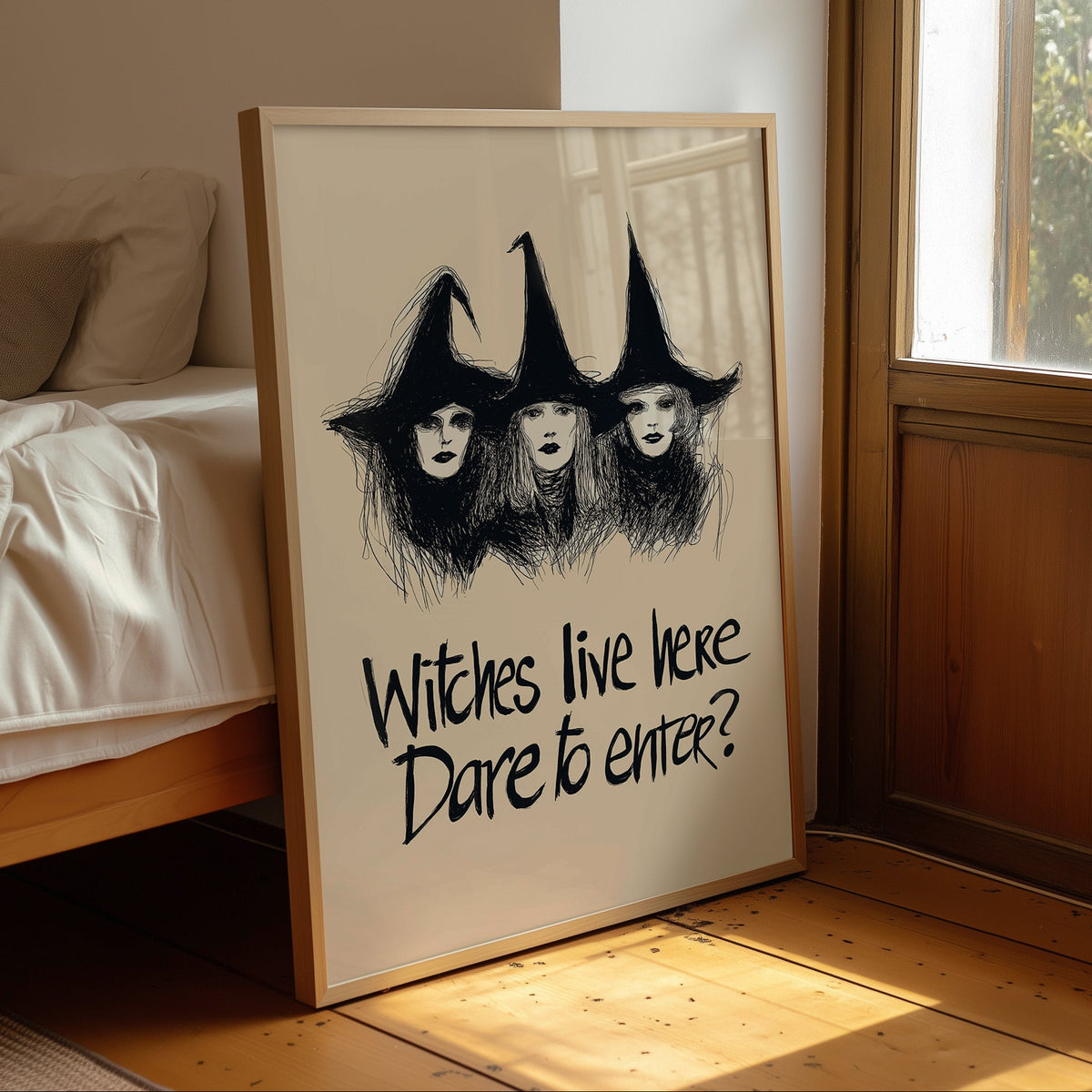 A framed art print leaning against a bed in a sunlit bedroom. The print shows three witch figures with tall pointed hats drawn in black sketch lines, with the text 'Witches live here.