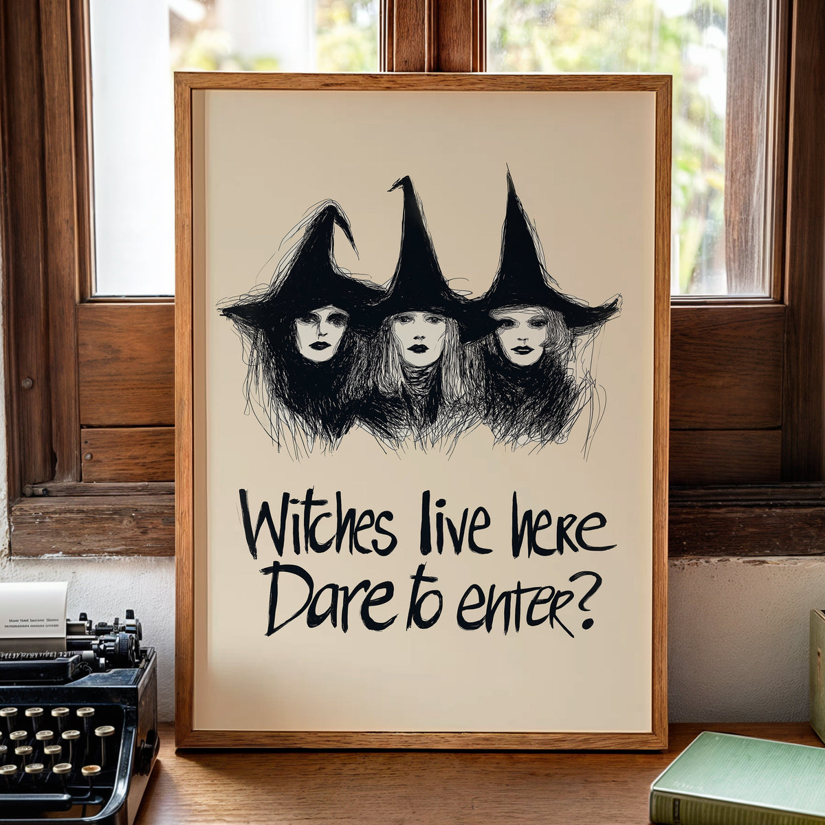 A framed art print featuring a black ink sketch of three witches with tall hats on an off-white background, with the text 'Witches live here Dare to enter?' below. The frame is set on a wooden table in a cozy room with a vintage typewriter, a book, and a candle nearby.