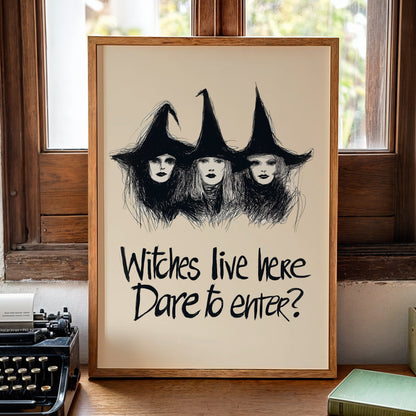 A framed art print featuring a black ink sketch of three witches with tall hats on an off-white background, with the text 'Witches live here Dare to enter?' below. The frame is set on a wooden table in a cozy room with a vintage typewriter, a book, and a candle nearby.