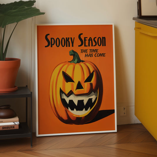 A framed vintage-style Halloween poster leaning against a white wall on a light wood floor. The poster features an orange background with the text 'Spooky Season' and 'The Time Has Come' above a carved orange pumpkin face.