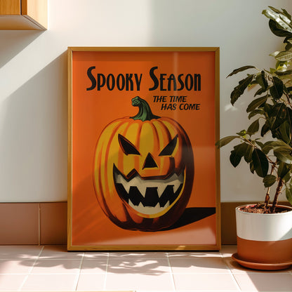 A framed vintage-style Halloween poster leaning against a wall on a tiled counter, next to a potted plant. The poster features an orange background with a carved jack-o'-lantern and the text 'Spooky Season' and 'The Time Has Come'.