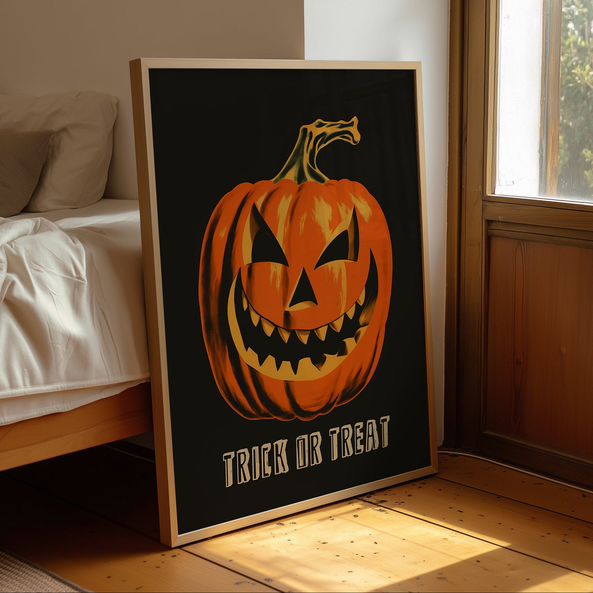 A framed Halloween art print leaning against a bed in a sunlit bedroom. The print shows a large orange jack-o'-lantern face on a black background with the words “TRICK OR TREAT” at the bottom.