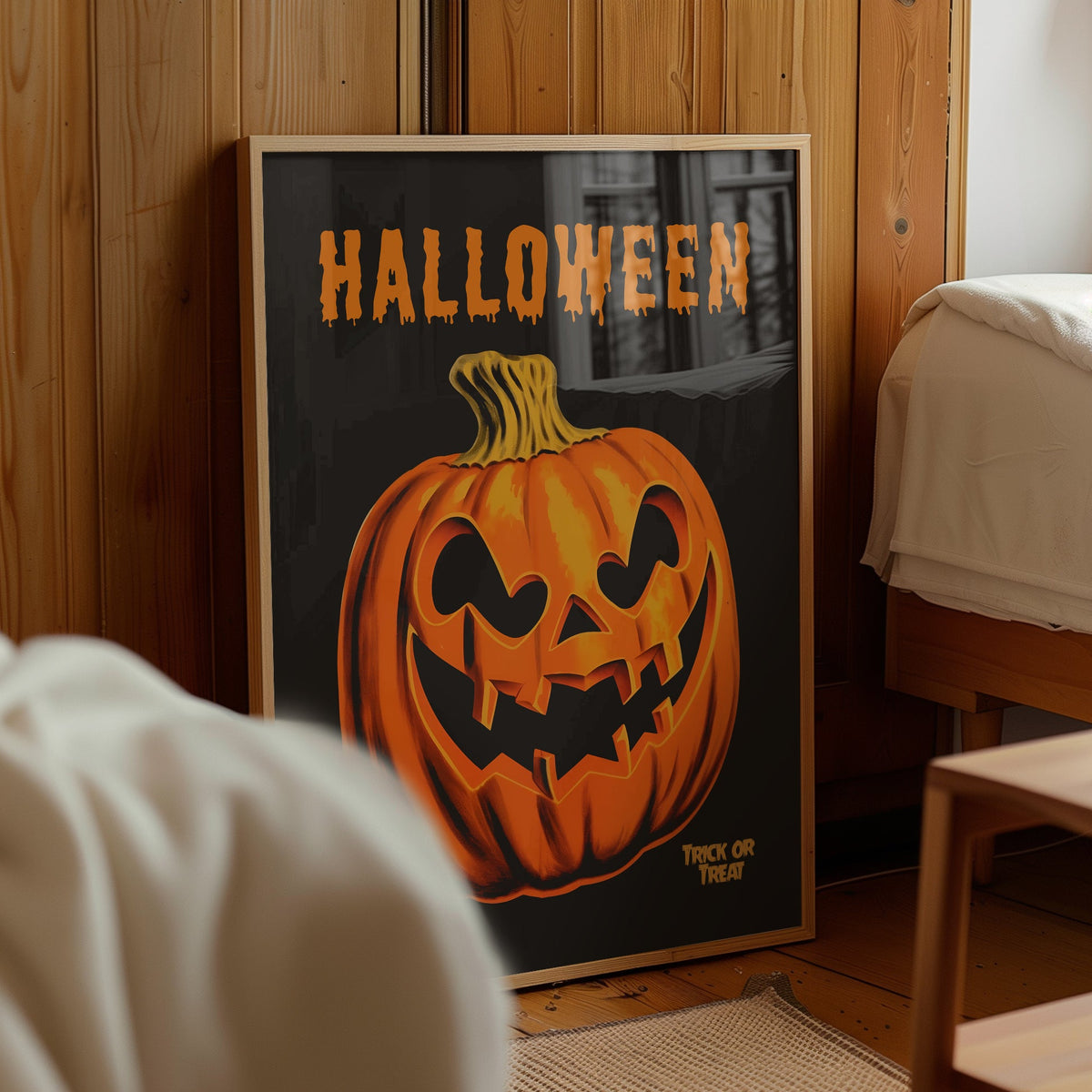 A framed Halloween art print leaning against a wooden wall in a bedroom. The print features a carved orange jack-o'-lantern pumpkin on a dark background with the word 'HALLOWEEN' in orange at the top and 'TRICK OR TREAT' at the bottom right.