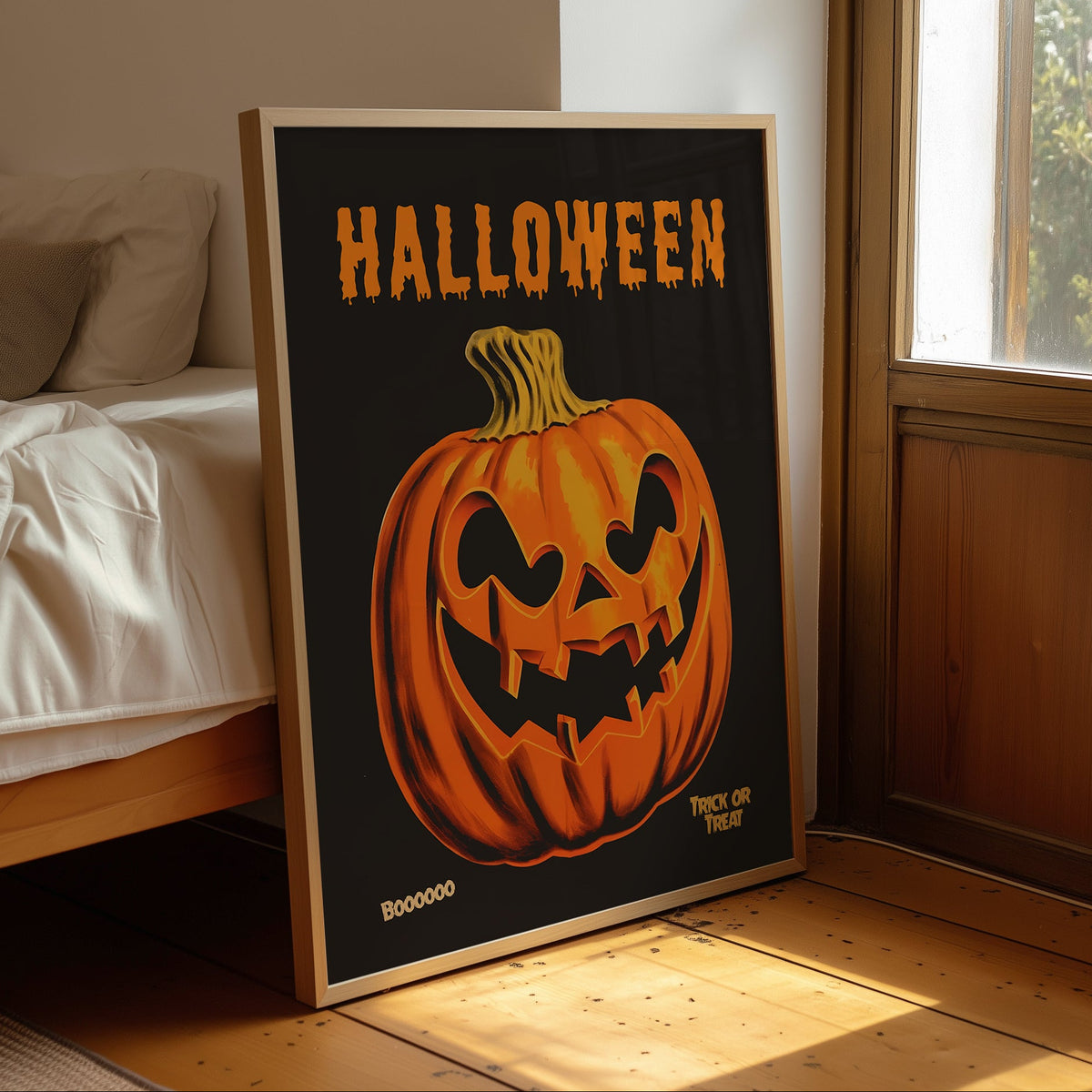 A framed Halloween art print leaning against a bed in a sunlit bedroom. The print features a large orange jack-o'-lantern on a black background with the word 'HALLOWEEN' in orange at the top.