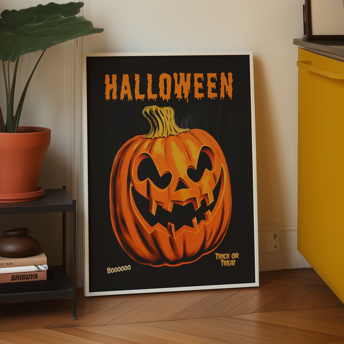 A framed Halloween art print leaning against a wall in a kitchen setting. The print features a large orange jack-o'-lantern pumpkin on a black background with the word 'HALLOWEEN' in orange at the top.