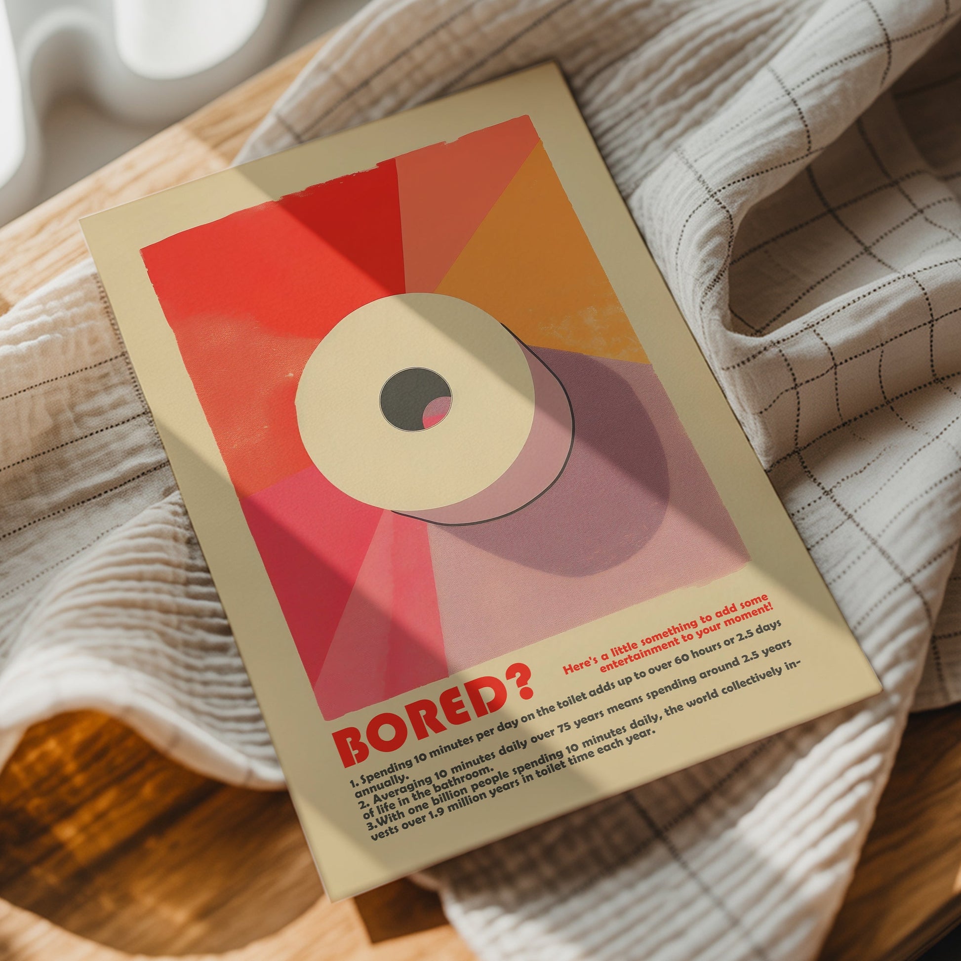 “Bored?” funny bathroom poster with a toilet roll illustration, styled on a beige checked blanket.