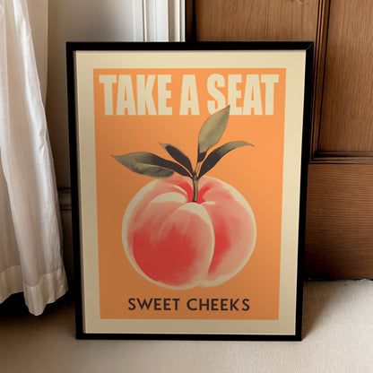 Framed Take a Seat Sweet Cheeks peach bathroom quote poster leaning beside a wooden cabinet and curtain.