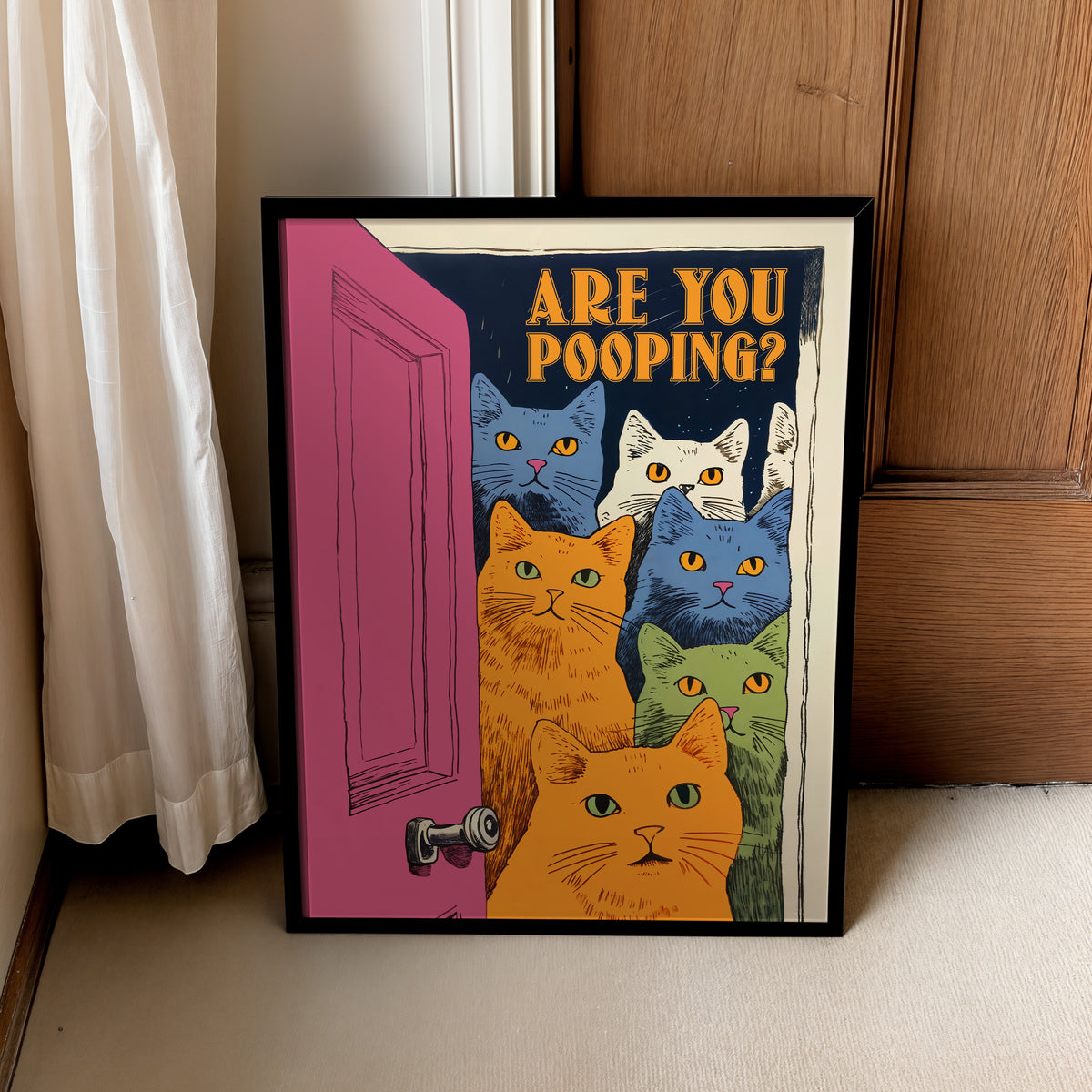 A framed art print leaning against a wooden door on a light-colored floor, next to a white curtain. The print features the text 'ARE YOU POOPING?' above a group of colorful cats peeking out from behind a pink door.
