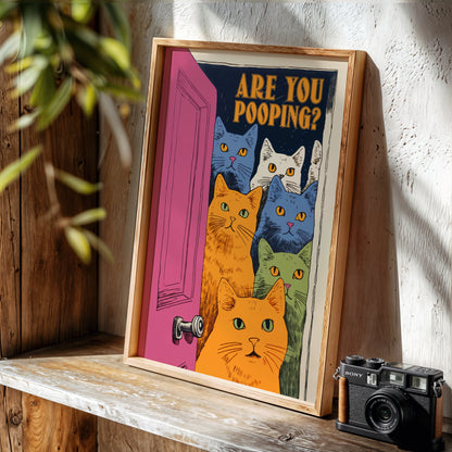 A framed art print leaning on a rustic wooden shelf next to a vintage Sony camera. The print features a humorous cartoon illustration of five cats peeking out from behind a pink door with the text 'ARE YOU POOPING?'.